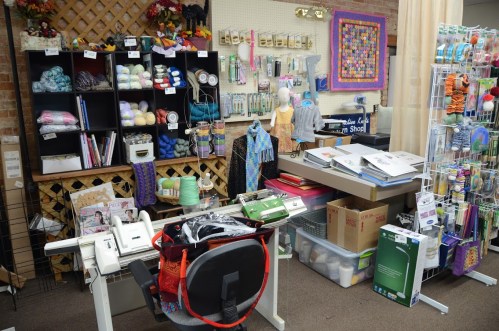 A cozy craft shop interior with shelves of yarn, tools, and supplies, plus a workspace with a chair and equipment.