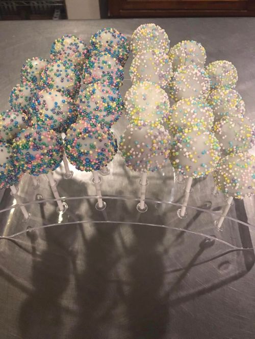 A display of cake pops coated in white chocolate and colorful sprinkles, arranged on a clear stand.