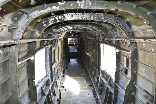 Interior view of an old aircraft fuselage, showing worn metal walls and empty space with sunlight streaming through windows.