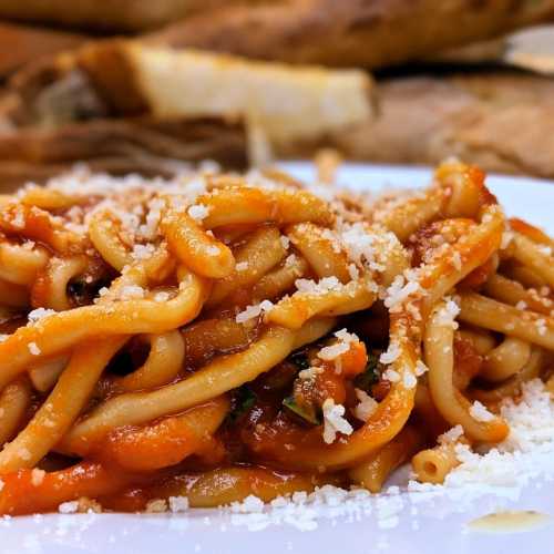 A close-up of pasta in tomato sauce, topped with grated cheese, served on a white plate with bread in the background.