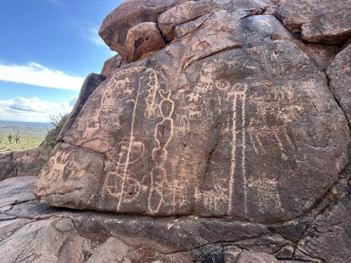 Petroglyphs carved into a rocky surface, depicting abstract figures and symbols against a desert landscape.