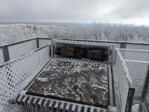 A frosty observation deck with a snow-covered map, surrounded by icy trees and a cloudy sky.