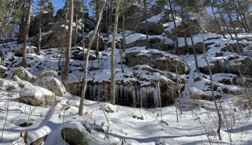 Snow-covered landscape with rocky outcrops and icicles hanging from the rocks, surrounded by tall trees.