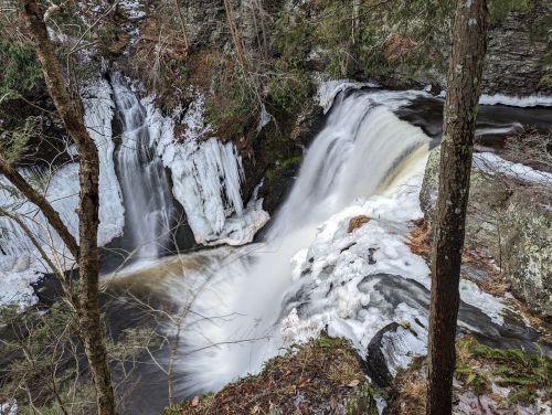 A serene waterfall cascades over icy rocks, surrounded by trees and winter scenery.