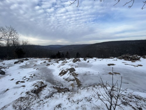 A snowy landscape with rocky terrain and a cloudy sky, showcasing distant hills and trees.
