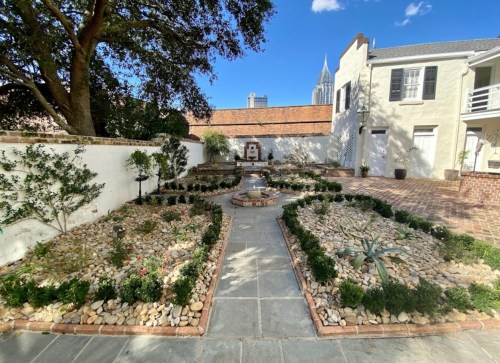 A landscaped garden with stone paths, plants, and a fountain, framed by buildings and a city skyline in the background.