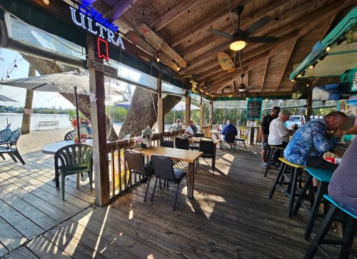 A rustic waterfront bar with wooden beams, tables, and patrons enjoying drinks under a sunny sky.