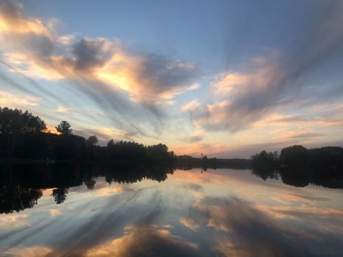 A serene lake at sunset, with colorful clouds reflecting on the water and trees lining the shore.