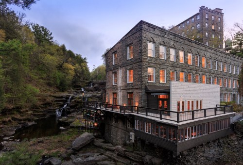 Historic stone building with large windows, overlooking a waterfall and surrounded by lush greenery at dusk.
