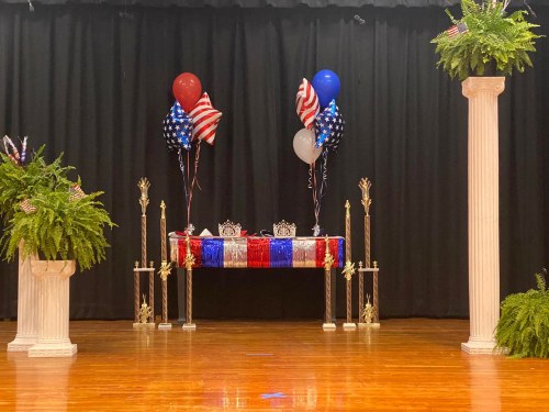 A decorated stage with a table draped in red, white, and blue, flanked by balloons and greenery, ready for an event.