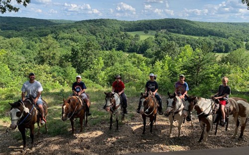 A group of six people on horseback, overlooking a lush green landscape with rolling hills and a blue sky.