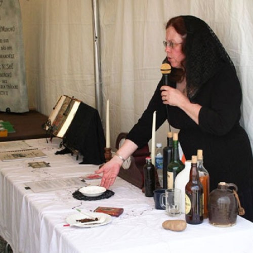 A woman in a black dress and veil speaks at a table with various bottles, candles, and documents in a tented setting.