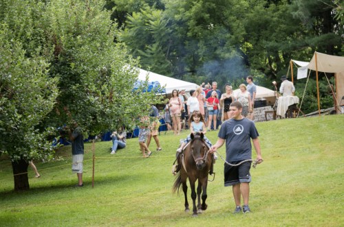 A child rides a small pony while being led by an adult, with a fair and people in the background on a grassy field.