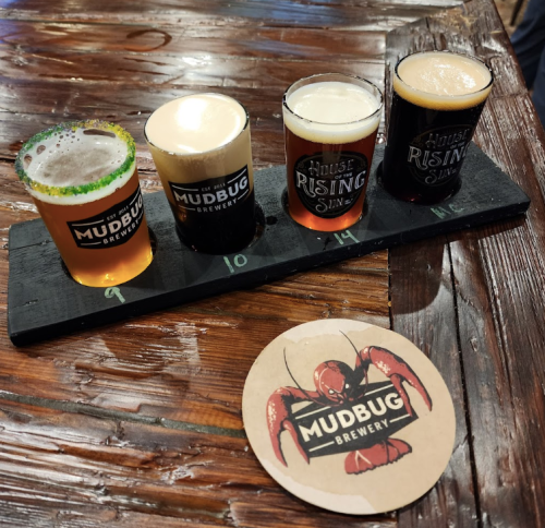Four small beer glasses on a wooden flight board, with a coaster featuring a lobster and the Mudbug Brewery logo.