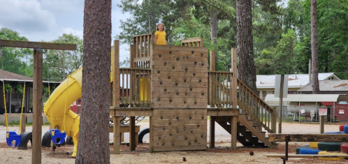A child in a yellow shirt stands on a wooden playground structure with a slide and climbing wall, surrounded by trees.