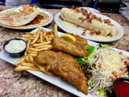 A plate of fried fish with fries and salad, alongside a large burrito and a taco platter in the background.