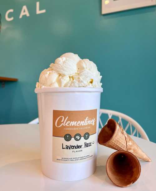 A container of lavender haze ice cream with a scoop on top, next to two empty ice cream cones, against a teal background.