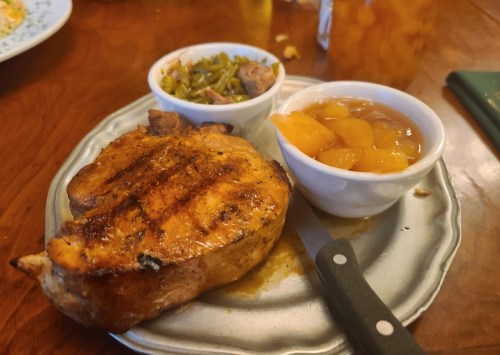A grilled pork chop on a plate, accompanied by bowls of green beans and peach slices.