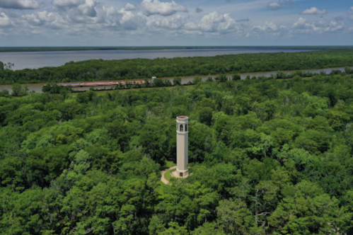 A tall, white tower stands amidst lush green trees, with a river visible in the background under a blue sky.