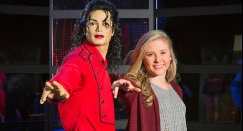 A girl poses next to a lifelike wax figure of a famous singer, both with arms outstretched, in a vibrant setting.