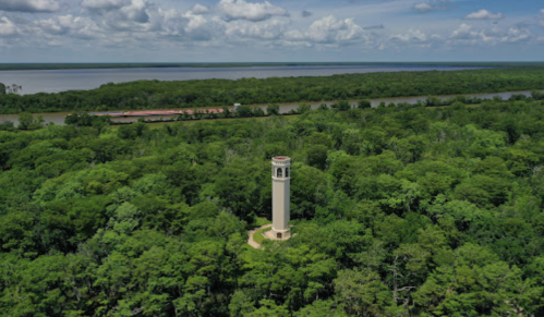 A tall tower stands amidst lush green trees, overlooking a river and a cloudy sky in the background.
