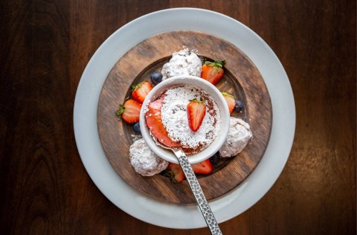 A bowl of dessert topped with powdered sugar and a strawberry, surrounded by fresh strawberries on a wooden platter.