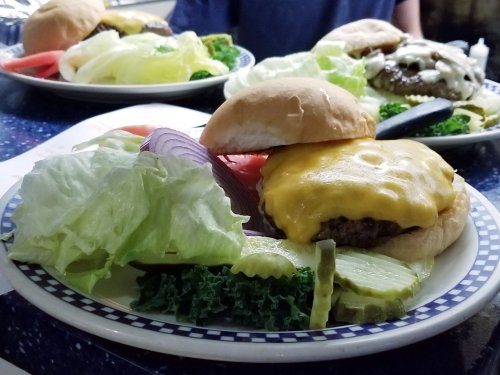 A cheeseburger with melted cheese, lettuce, tomato, pickles, and onion on a plate, accompanied by a side of greens.