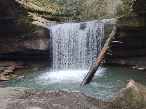 A serene waterfall cascades over rocky cliffs into a tranquil pool, surrounded by lush greenery.