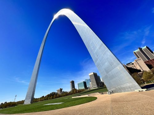 A tall, shiny arch structure against a clear blue sky, surrounded by green grass and modern buildings.