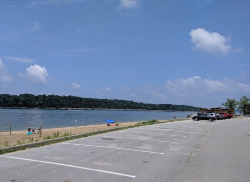 A sandy beach by a river, with a few people enjoying the water and parked cars along the shore under a blue sky.