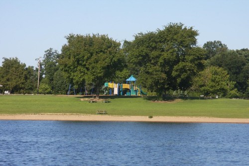 A playground with colorful equipment surrounded by trees, viewed across a calm lake on a clear day.
