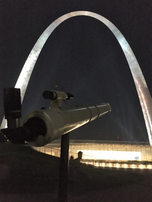 A telescope in the foreground with the Gateway Arch illuminated at night in the background.
