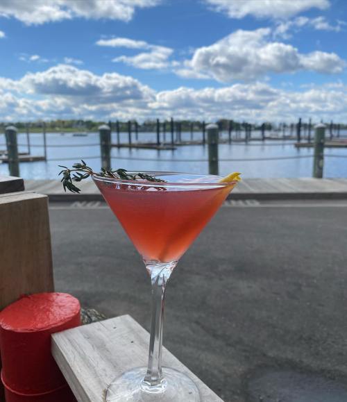 A vibrant cocktail garnished with thyme and lemon, set against a scenic waterfront with clouds in the sky.