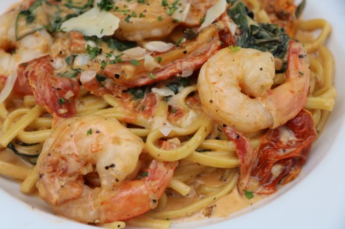 A close-up of shrimp pasta with creamy sauce, sun-dried tomatoes, spinach, and garnished with herbs and cheese.