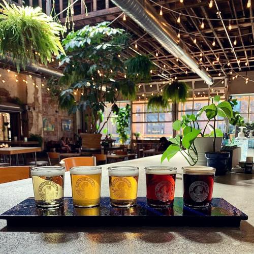 A wooden tray holds five glasses of different colored drinks, surrounded by greenery and warm string lights in a cozy setting.