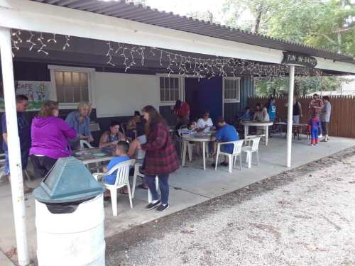 A group of people gathered outside under a covered area, engaging in activities at tables. Trees and a fence are in the background.
