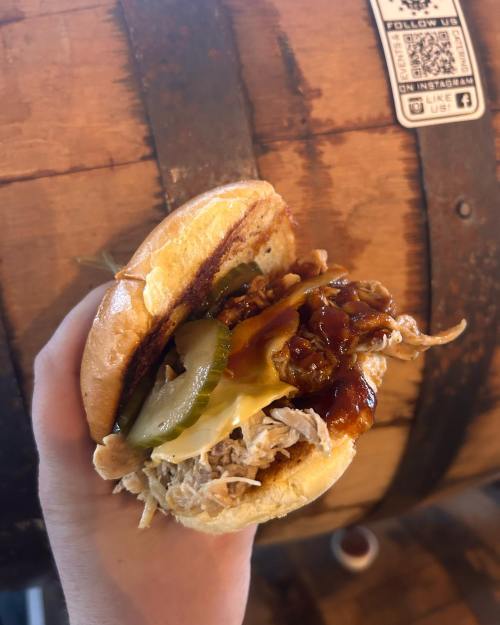 A hand holding a pulled pork sandwich with pickles and barbecue sauce, set against a wooden barrel background.