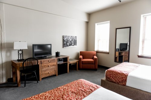 A cozy hotel room featuring two beds, a TV, a chair, and a wooden desk with a lamp and mirror. Natural light from windows.