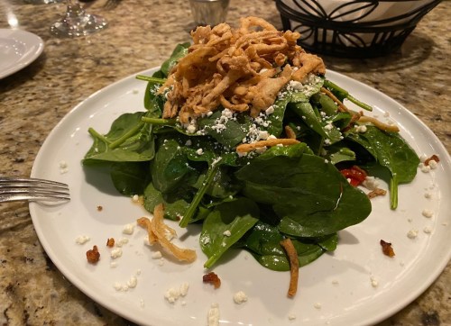A fresh spinach salad topped with crispy onions and crumbled cheese, served on a white plate.