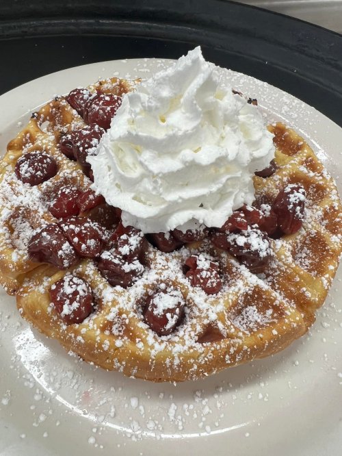 A golden waffle topped with whipped cream and cherries, dusted with powdered sugar on a white plate.