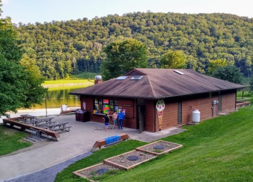 A wooden building by a lake, with picnic tables and people outside, surrounded by green hills and trees.