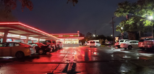A diner with neon lights at night, reflecting on a wet parking lot filled with cars.