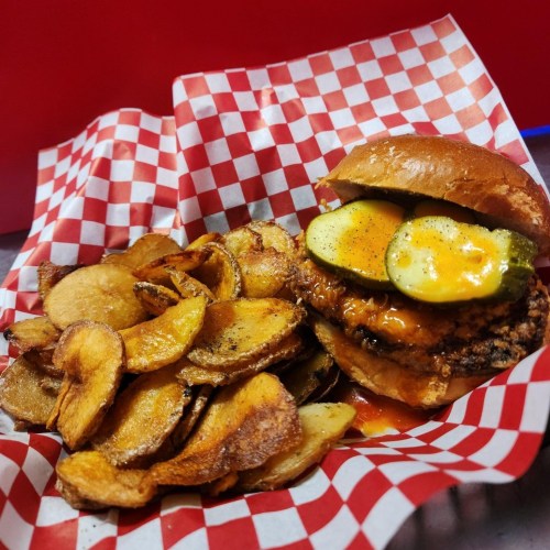 A crispy chicken sandwich with pickles and sauce, served with a side of golden, crispy potato chips in a red checkered basket.