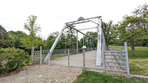 A metal truss bridge spans a grassy area, surrounded by trees under a cloudy sky.