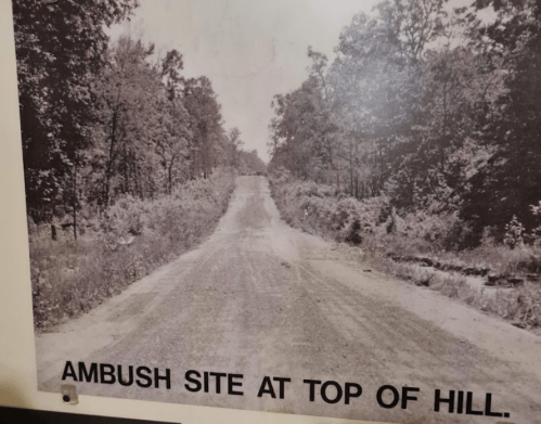 Black and white photo of a dirt road lined with trees, labeled "Ambush Site at Top of Hill."