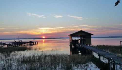 Sunset over a calm lake, with a wooden dock and a small building on the shore, surrounded by tall grass.