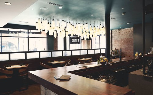A modern restaurant interior featuring a bar, stylish lighting, and large windows with a view of the street outside.