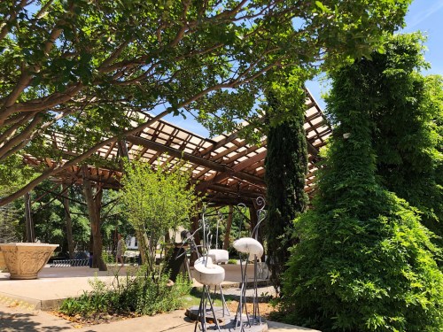 A serene outdoor space with a wooden pergola, lush greenery, and decorative sculptures under a clear blue sky.