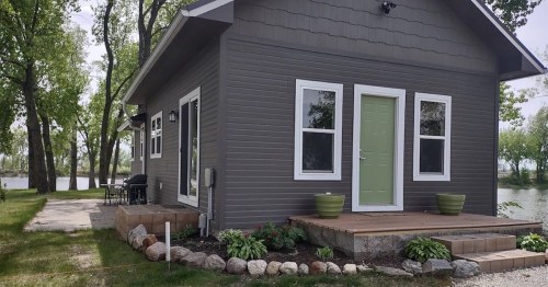 A modern gray house with green door, surrounded by trees and a stone pathway near a body of water.