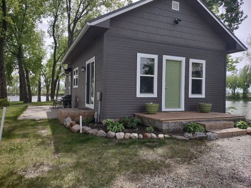 A modern gray cottage with a green door, surrounded by greenery and a stone pathway near a calm body of water.